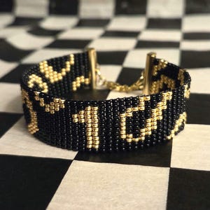 Könnte beinhalten: Schwarzes und goldenes Perlenarmband mit geometrischem Muster. Das Armband besteht aus kleinen, runden Perlen und hat einen goldenen Verschluss und eine Kette. Das Armband ist etwa 2,5 cm breit.