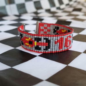 Puede incluir: Un brazalete de cuentas rojas, grises y negras con un patrón a cuadros y un pequeño diseño de corazón rojo. El brazalete está hecho de pequeñas cuentas redondas y tiene un cierre plateado.