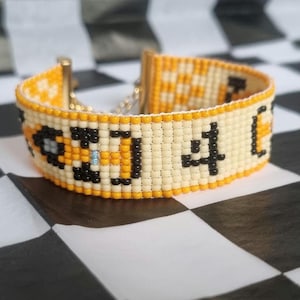 Puede incluir: Un brazalete de cuentas blancas y amarillas con cuentas negras formando un patrón. El brazalete tiene un cierre dorado.