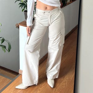 May include: A white long-sleeve top with a knot detail paired with cream-colored cargo pants. The pants have side pockets and a zipper detail. The outfit is completed with white pointed-toe shoes. The mirror is black and round.