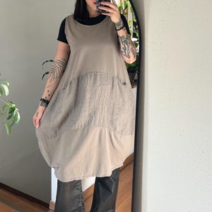 May include: A sleeveless, beige linen dress with a large front pocket and a rounded hem. The dress is layered over a black top and paired with black pants. The image is reflected in a large, oval mirror.