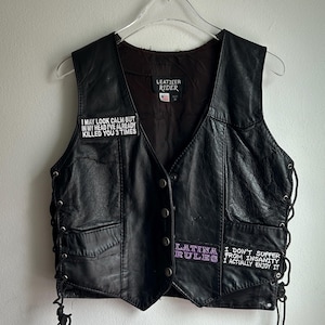 Op de afbeelding: Zwarte leren vest met een knoopsluiting, veter aan de zijkant en twee voorzakken. Patches met tekst zijn onder meer "I MAY LOOK CALM BUT IN MY HEAD I'VE ALREADY KILLED YOU 3 TIMES", "LATINA RULES" en "I DON'T SUFFER FROM INSANITY I ACTUALLY ENJOY IT".
