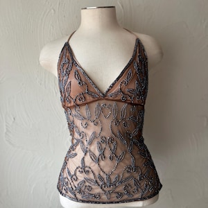 Puede incluir: Un top halter sin mangas hecho de tela de malla transparente color carne con intrincados abalorios gris oscuro en un patrón de hojas y enredaderas. La prenda tiene un escote en V profundo, tirantes finos y una silueta ajustada hasta la cintura.