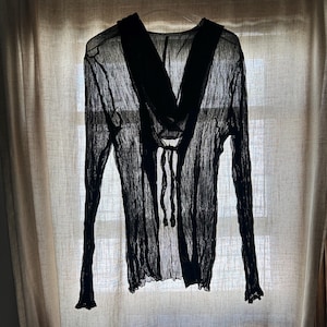 May include: A black long-sleeved shirt made of a sheer, crinkled fabric. The garment features a wide collar, a front opening with a tie closure at the chest, and a button-down placket extending toward the hem. The sleeves are long with ruffled cuffs.