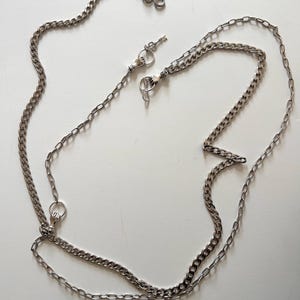 May include: A silver-colored chain with a small key charm and clasp. The chain is made of interlocking links and is designed to be worn as a necklace or used for other decorative purposes. The chain is displayed on a white surface.