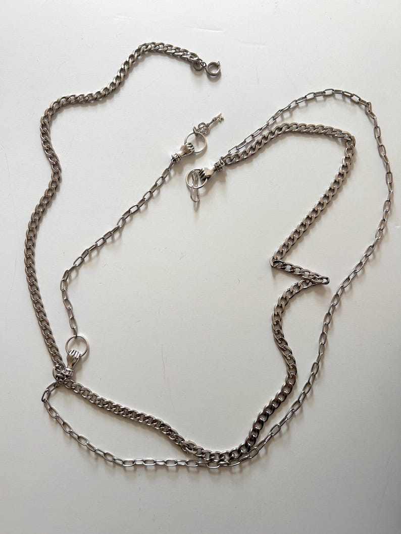 May include: A silver-colored chain with a small key charm and clasp. The chain is made of interlocking links and is designed to be worn as a necklace or used for other decorative purposes. The chain is displayed on a white surface.
