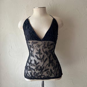 May include: A sleeveless black top featuring a deep V-neckline and thin halter straps. The garment is crafted from a sheer, nude-coloured mesh fabric adorned with intricate black beadwork in a swirling vine and leaf pattern throughout the bodice.
