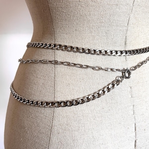 May include: Silver-toned chain belt with three strands of varying chain link sizes. The belt is displayed on a mannequin, showcasing the waist-level placement and the different chain styles. The chains are connected with a clasp.