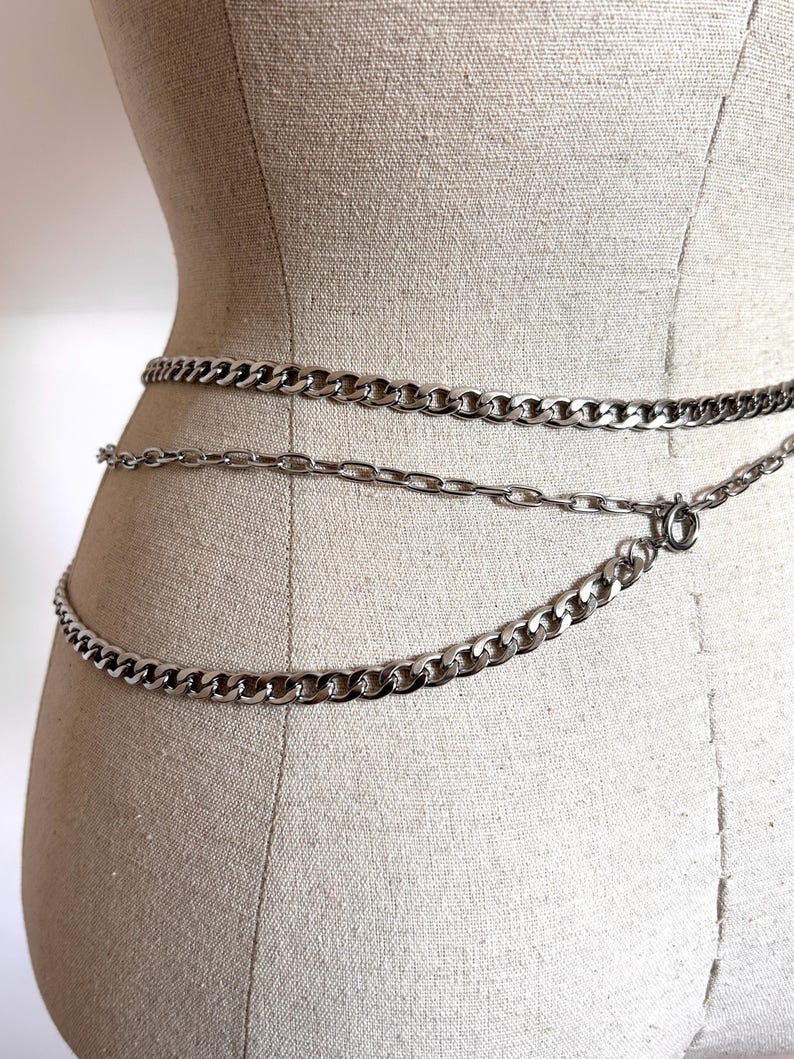 May include: Silver-toned chain belt with three strands of varying chain link sizes. The belt is displayed on a mannequin, showcasing the waist-level placement and the different chain styles. The chains are connected with a clasp.