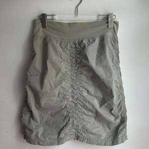 May include: A light olive green skirt hangs from a silver hanger against a white wall. The skirt has a high waist and a gathered, ruched design down the center. The fabric appears lightweight and slightly textured, with a subtle sheen.