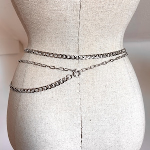 May include: Silver-toned chain belt with three strands of varying chain link sizes. The belt is fastened with a circular clasp. The belt is displayed on a mannequin.