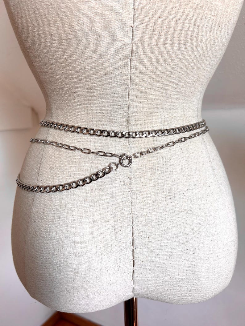 May include: Silver-toned chain belt with three strands of varying chain link sizes. The belt is fastened with a circular clasp. The belt is displayed on a mannequin.