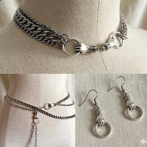 May include: A silver-toned jewelry set featuring a choker necklace, a waist chain, and a pair of earrings. The necklace and earrings have a hand holding a ring design. The waist chain has a dangling chain.