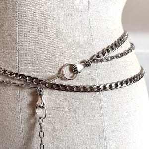 May include: Silver chain belt featuring a hand-shaped clasp and ring detail. The belt is made up of multiple chain strands and is shown on a mannequin. The chains are linked and have a polished finish.