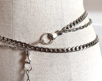 May include: Silver-toned chain belt with a hand-shaped clasp and ring detail. The belt is composed of multiple chain strands and is displayed on a mannequin. The chains are linked together and have a shiny finish.