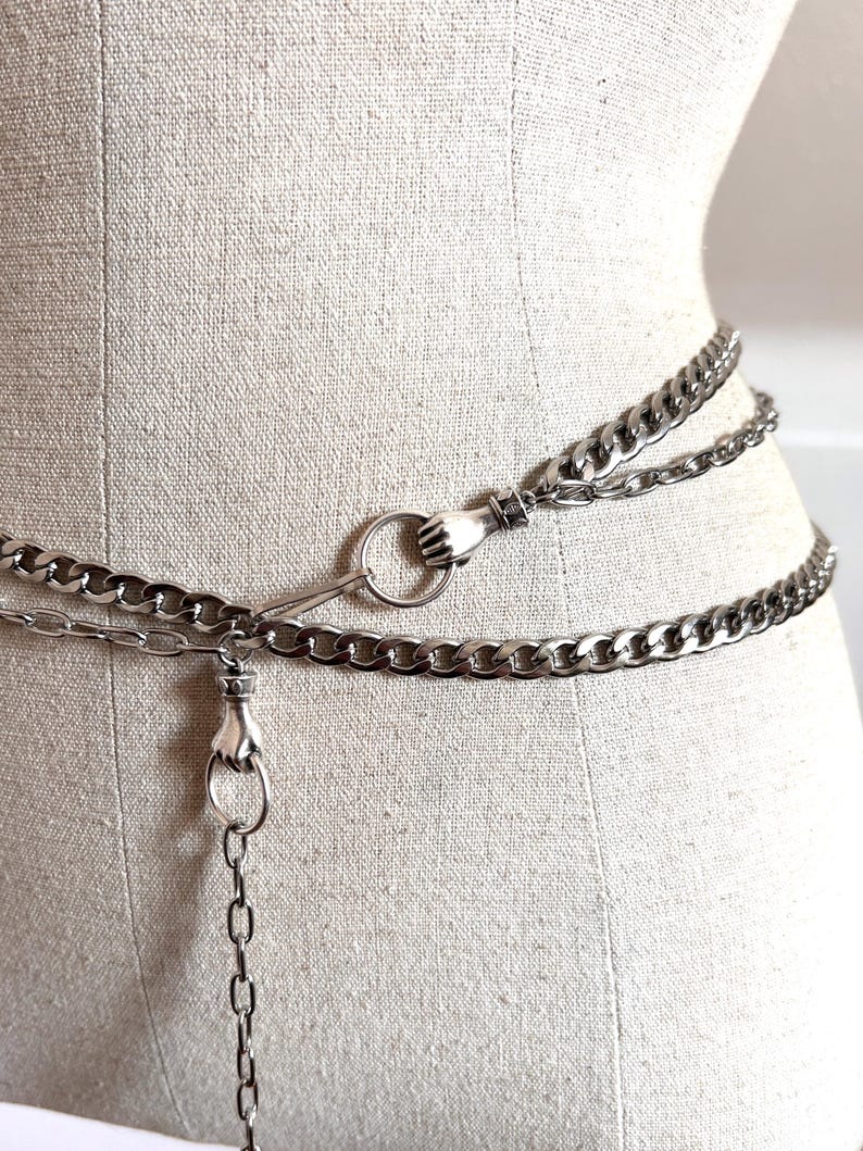 May include: Silver-toned chain belt with a hand-shaped clasp and ring detail. The belt is composed of multiple chain strands and is displayed on a mannequin. The chains are linked together and have a shiny finish.