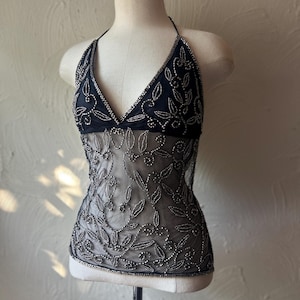 May include: A black halter top featuring a sheer mesh midsection and bodice adorned with intricate silver beadwork in a floral vine pattern. The garment has a deep V-neckline, thin halter straps, and a fitted silhouette that extends to the waist.