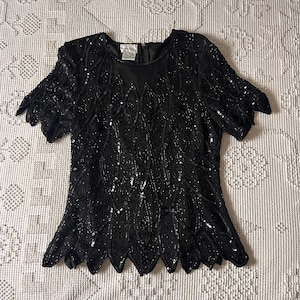 May include: A black short-sleeved top featuring an intricate pattern of black sequins and beads arranged in leaf-like shapes. The garment has a round neckline, a back zip closure, and a scalloped hemline. A label reads "Barrie Pace".