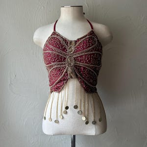 May include: A deep red halter crop top featuring intricate gold beadwork in a butterfly-inspired pattern across the front. The garment includes thin straps and a series of dangling gold chains finished with small circular metal coins at the hem.
