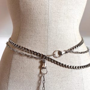 May include: Silver-toned chain belt with three layers of chains. The belt features a hand-shaped clasp and a hand-shaped ring detail. The belt is displayed on a mannequin.