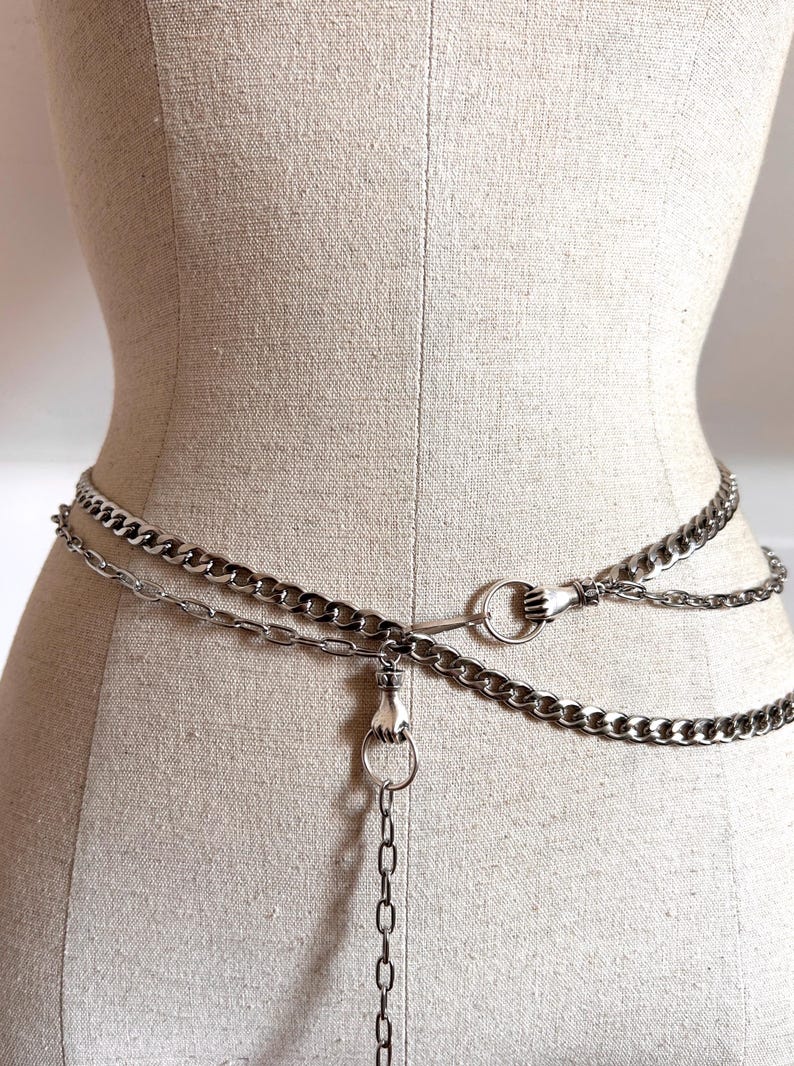 May include: Silver-toned chain belt with three layers of chains. The belt features a hand-shaped clasp and a hand-shaped ring detail. The belt is displayed on a mannequin.