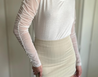 Könnte beinhalten: Ein weißes Langarmshirt mit durchsichtigen Mesh-Ärmeln und einem beigefarbenen gerippten Minirock.