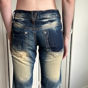 May include: Blue denim jeans with a faded, distressed design. The jeans feature a light beige wash on the front and back, with darker blue accents. The back pockets have a unique shape and button details. The jeans have a low waist and a straight leg.