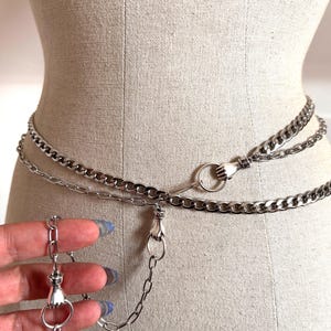 May include: Silver chain belt with three strands, featuring hand-shaped clasps and a key pendant. The belt is displayed on a mannequin, showcasing its design and style. The chain links are medium-sized and the overall aesthetic is gothic.