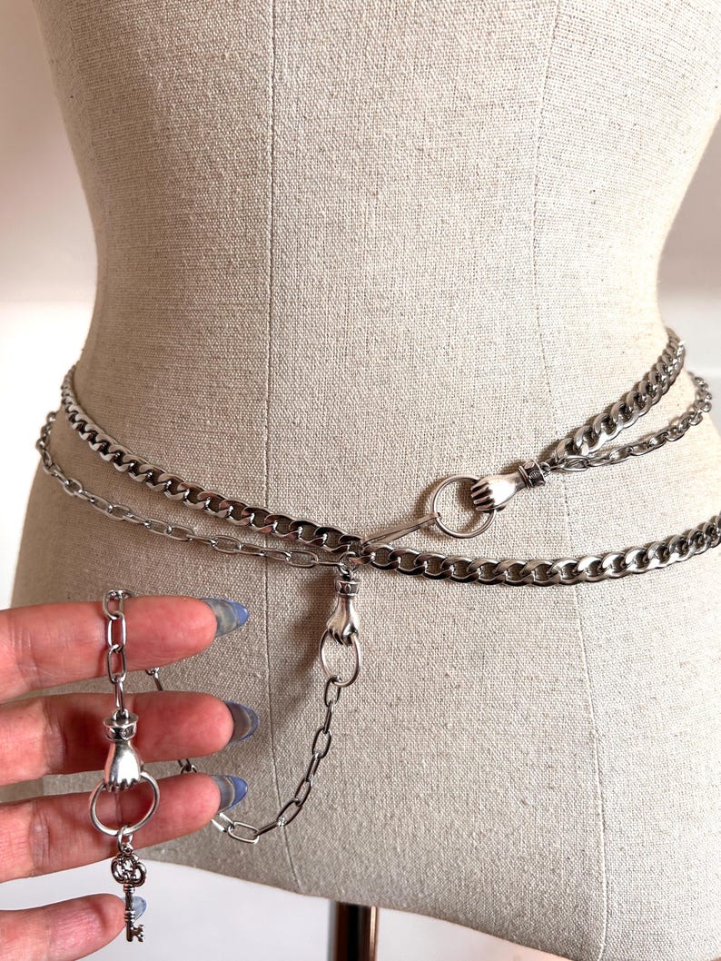 May include: Silver chain belt with three strands, featuring hand-shaped clasps and a key pendant. The belt is displayed on a mannequin, showcasing its design and style. The chain links are medium-sized and the overall aesthetic is gothic.