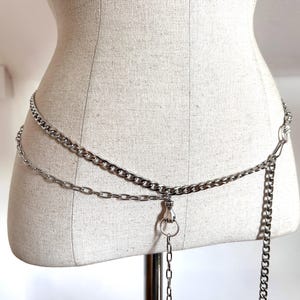 May include: Silver-toned chain belt with two parallel chains and a dangling chain. The belt features a clasp and a ring detail. The chains are displayed on a mannequin.