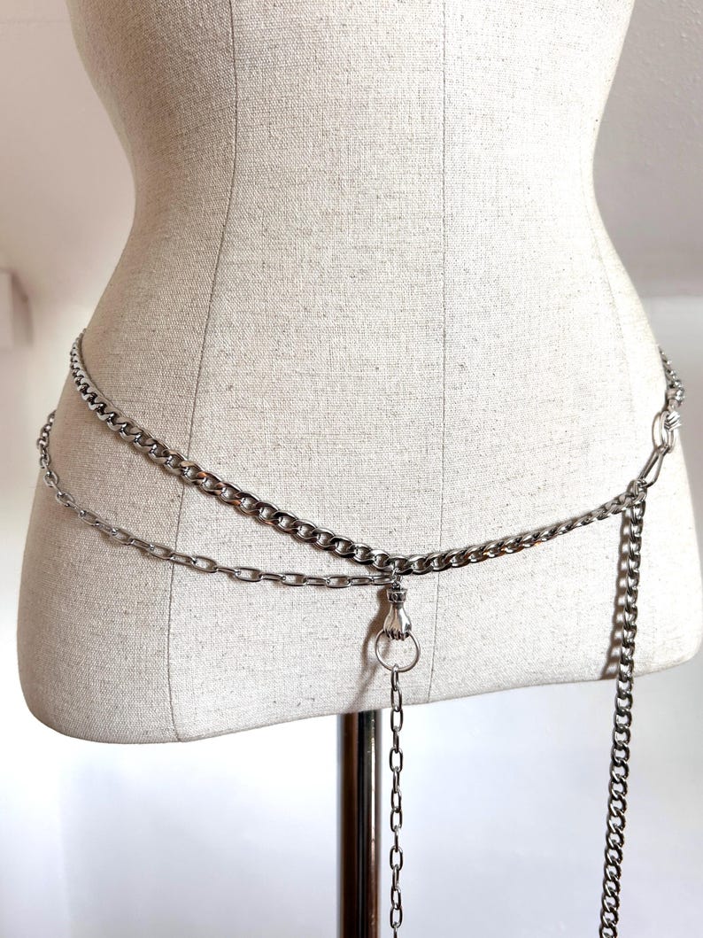 May include: Silver-toned chain belt with two parallel chains and a dangling chain. The belt features a clasp and a ring detail. The chains are displayed on a mannequin.