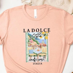 a pink t-shirt with a graphic design of a coastal scene, along with the text "LADOLCE VITA ANTIGIO ITALIA". The t-shirt is displayed on a white background, surrounded by various accessories such as a straw hat, a woven basket, and a potted plant.
