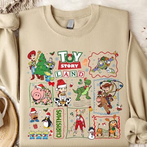 a beige T-shirt with a collage of various cartoon characters and scenes from the Toy Story Land movie franchise, including characters like Woody, Buzz Lightyear, and a snowman. The T-shirt is displayed on a bed with a white blanket and a mug, creating a cozy and festive atmosphere.