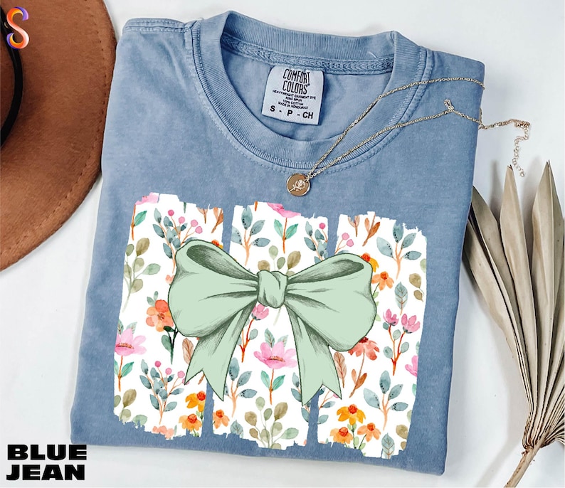 A blue denim t-shirt with a bow design and a floral pattern, along with a necklace and some dried flowers, are displayed on a white surface.