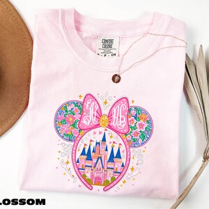 A pink t-shirt with a colorful illustration of a castle and flowers, along with a pink bow on the head. The t-shirt is placed on a white surface, and there is a brown hat and a plant in the background.