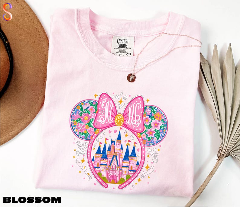 A pink t-shirt with a colorful illustration of a castle and flowers, along with a pink bow on the head. The t-shirt is placed on a white surface, and there is a brown hat and a plant in the background.