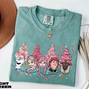 A light green t-shirt with a Christmas tree design and characters from the Disney movie Frozen, including Olaf, Elsa, Anna, and a reindeer.
