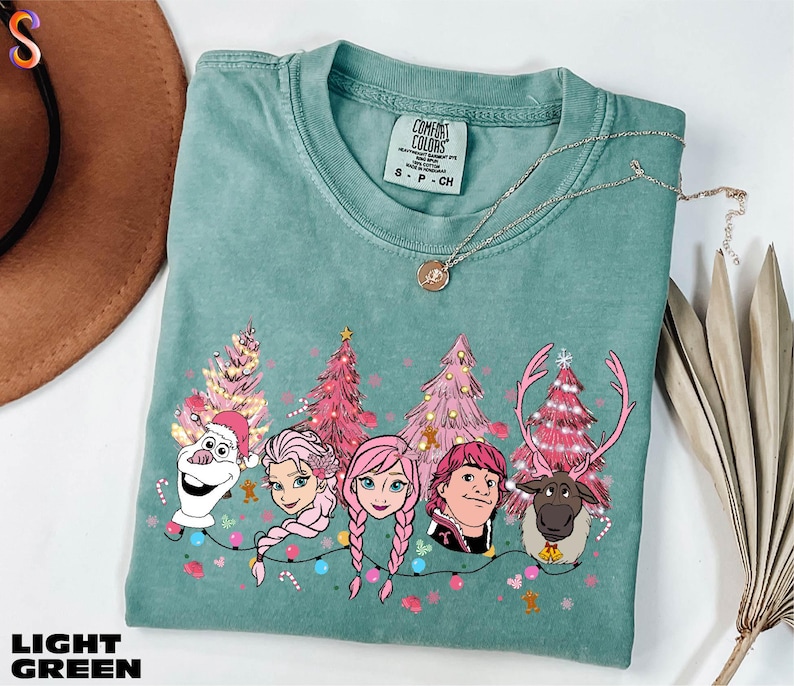 A light green t-shirt with a Christmas tree design and characters from the Disney movie Frozen, including Olaf, Elsa, Anna, and a reindeer.