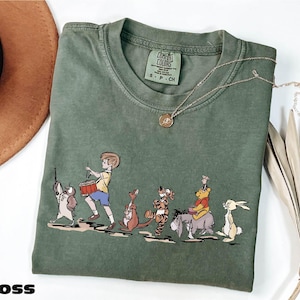 May include: Moss green t-shirt featuring a cartoon illustration of Winnie the Pooh and friends. The design includes characters like Piglet, Pooh, and Tigger. The shirt has a crew neck and short sleeves. A gold necklace is draped across the shirt.
