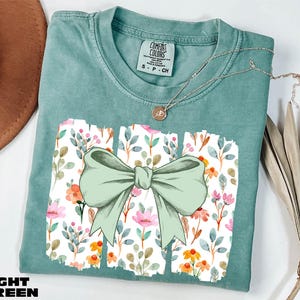 A light green t-shirt with a bow design and a floral pattern, along with a necklace and some dried leaves, are displayed on a white surface.