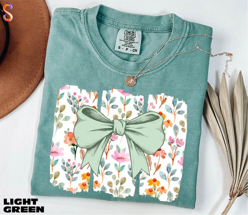 A light green t-shirt with a bow design and a floral pattern, along with a necklace and some dried leaves, are displayed on a white surface.