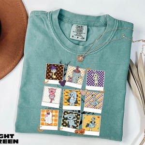 A light green t-shirt with a graphic design of various cartoon characters, including a ghost, a skeleton, and a pumpkin, is displayed on a white surface alongside a brown hat and some dried leaves.