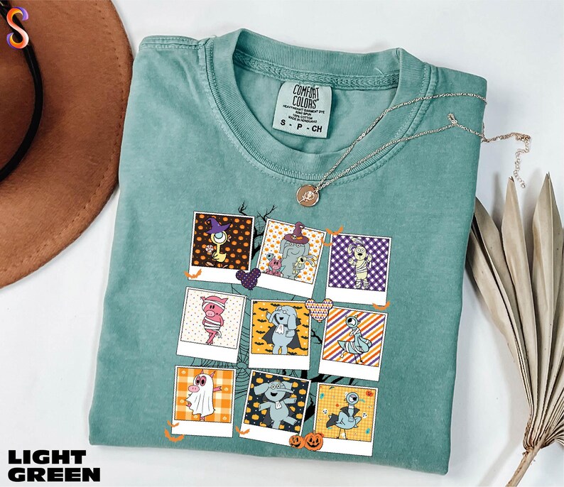 A light green t-shirt with a graphic design of various cartoon characters, including a ghost, a skeleton, and a pumpkin, is displayed on a white surface alongside a brown hat and some dried leaves.