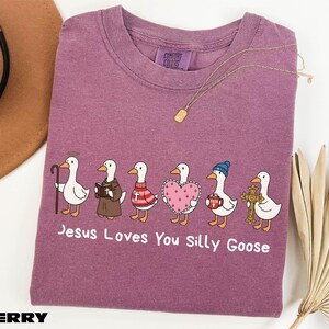 A purple t-shirt with a graphic design of six cartoon ducks, each with a different outfit or accessory, and the text "Jesus Loves You Silly Goose" printed on it.