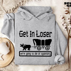 May include: Sport grey hoodie with the text "Get In Loser" in a pixelated font, along with a black wagon and ox graphic. The phrase "we're going to die of dysentery" is below the graphic. The hoodie has a drawstring hood.