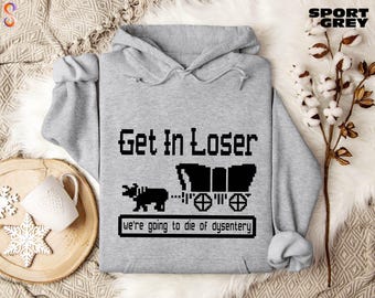 May include: Sport grey hoodie with the text "Get In Loser" in a pixelated font, along with a black wagon and ox graphic. The phrase "we're going to die of dysentery" is below the graphic. The hoodie has a drawstring hood.