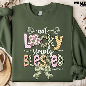 Puede incluir: Sudadera verde militar con el texto "not lucky simply blessed" en un diseño colorido y estampado. El diseño incluye tréboles, lunares y un lazo. También se incluye el texto "Romanos 4:7-8".