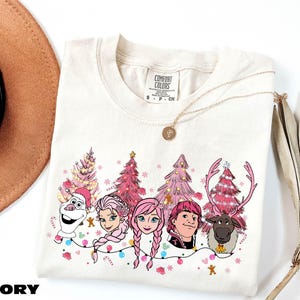 A white t-shirt with a graphic design featuring characters from the Disney movie Frozen, including Olaf, Elsa, Anna, and a reindeer. The characters are surrounded by Christmas trees and decorations, creating a festive and whimsical scene.