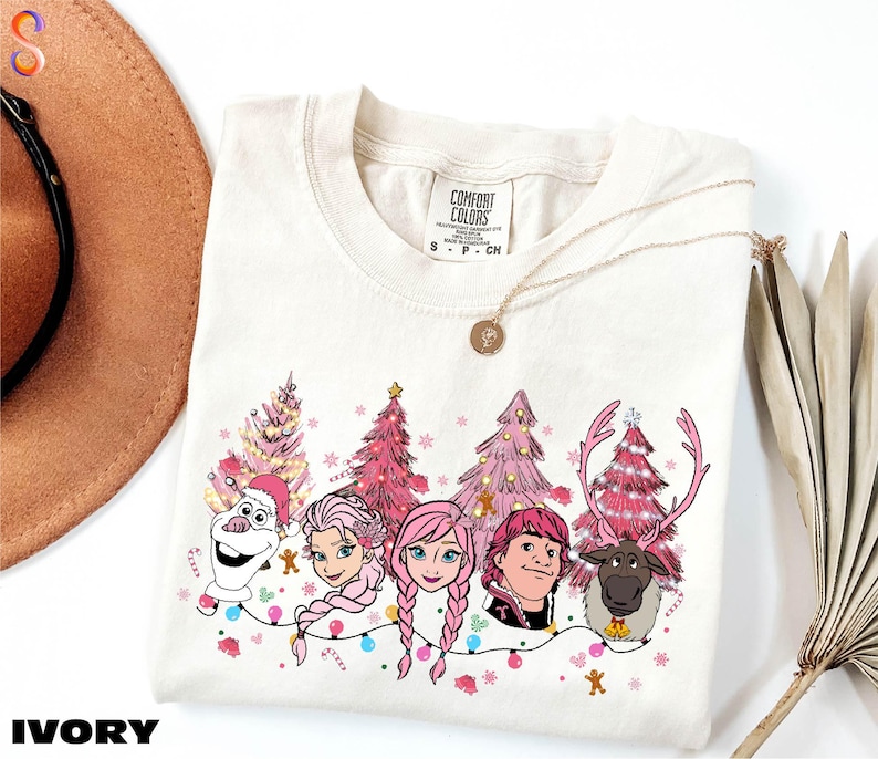 A white t-shirt with a graphic design featuring characters from the Disney movie Frozen, including Olaf, Elsa, Anna, and a reindeer. The characters are surrounded by Christmas trees and decorations, creating a festive and whimsical scene.