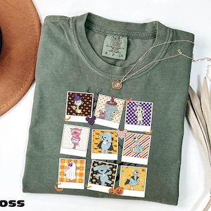 A green t-shirt with a graphic design of various cartoon characters and illustrations, including a witch, pumpkin, and other Halloween-themed elements. The t-shirt is folded and placed on a white surface, with a brown hat and some dried leaves nearby.
